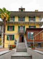 A two-story yellow building with green shutters and balconies, featuring a staircase leading to the entrance and a small wooden annex nearby.