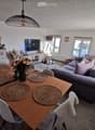 Modern living and dining area with a grey sofa, wooden dining table with six chairs, a television, and decorative items including a flower vase and a floor lamp.