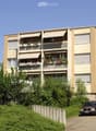 Four-story residential building with balconies, awnings, and flower boxes, surrounded by greenery and a parking area.