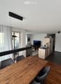 Open-plan living and dining area with a wooden dining table, chairs, a television, a small kitchen area with a sink, refrigerator, and a balcony with sheer curtains.