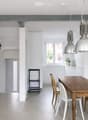 Bright, open-plan dining area with a wooden dining table, white and wooden chairs, pendant lights, and a small stool near the kitchen area.