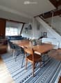 Bright living and dining area with wooden floors, a large dining table with chairs, a sofa, a staircase, and modern decor including a patterned rug and pendant lights.