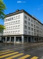 Multi-story building with white facade, multiple windows, and a flat roof, located on a street with tram tracks and a bus lane.