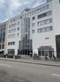 Modern multi-story building with white facade, large windows, and a cylindrical tower section. The ground floor has glass storefronts with signage reading "GENERALI" and "WEIGTIKLASSE 11". Bicycles are parked along the sidewalk in front of the building.