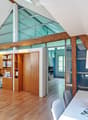 Bright, modern room with wooden flooring, large glass windows, and exposed wooden beams. Features a white table with a potted plant, chairs, a wooden cabinet with shelves, and a desk area with a telephone and books.