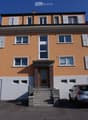 Two-story house with orange facade, white shutters, and a central entrance with steps. Features two garage doors and a small balcony on the upper floor.