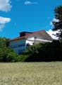 Two-story house with a red-tiled roof, white exterior, and a balcony on the upper floor, surrounded by trees and a grassy field.