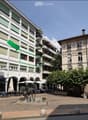 Multi-story building with balconies and green shutters, located in an urban area with outdoor seating, a fountain, and trees.