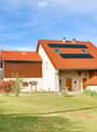 Two-story house with red-tiled roof, solar panels, stone and wood exterior, surrounded by lawn and trees.
