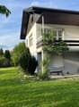 Two-story house with a modern design, featuring a balcony on the upper level, surrounded by a well-maintained lawn and trees.