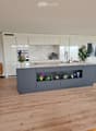 Modern kitchen with a grey island, white cabinets, marble backsplash, and wooden flooring. Features a coffee maker, sink, and decorative plants on the island and shelves.