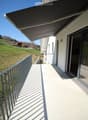 Balcony with a metal railing, covered by a retractable awning, overlooking a green hillside with scattered houses.