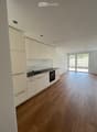 Empty kitchen with light-colored cabinets, a sink, and a built-in oven. The floor is made of light wood, and there is a large window with blinds leading to an outdoor area.
