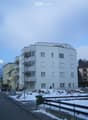 Multi-story residential building with balconies and windows, surrounded by other buildings and a snow-covered landscape.