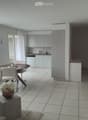 Bright, modern room with tiled flooring, a round wooden dining table with four white chairs, a vase with white flowers on a small table, and a kitchen area with white cabinets and a sink.