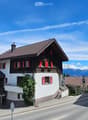House. Traditional Swiss house with red shutters, white walls, and a tiled roof, situated in a scenic mountain landscape.
