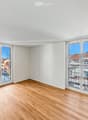 Empty room with light wooden flooring and white walls, featuring large sliding glass doors that open to a balcony with a view of rooftops.
