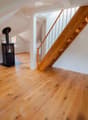 Empty attic space with wooden flooring, white walls, and a staircase leading upwards. A black wood-burning stove is positioned against the wall.