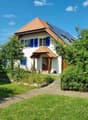 Two-story house with white walls, blue shutters, red-tiled roof, and solar panels on the roof. The house has a small porch with a wooden door and is surrounded by a well-maintained garden with a gravel pathway and wooden fence.