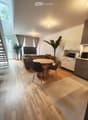 Open-plan living and dining area with a sofa, dining table, chairs, and a television. Kitchen area features white cabinets and a microwave. Wooden flooring throughout.