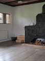 Bright room with wooden flooring, white walls, and a large wood stove. Features include a radiator, wicker basket, wooden crate, teapot, and a stack of firewood.