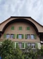 Two-story house with brown tiled roof and green shutters, surrounded by trees and a small garden area.