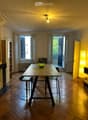 Dining area with wooden herringbone floor, modern table with four stools, green bowl on table, large windows with shutters, warm lighting from pendant lamp and floor lamp.