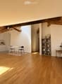 Open-plan living space with a bedroom and kitchen area. Features a wooden floor, exposed wooden beams, a grey upholstered bed, a nightstand, a small table with chairs, a kitchen with white cabinets, an oven, and a shelf with decorative items.
