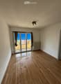 Empty room with wooden flooring, white walls, and a large sliding glass door leading to a balcony. The room has a ceiling light fixture and a radiator on one wall.