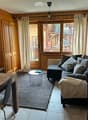 A cozy living room with wooden furniture, a sofa, coffee table, and a dining table. Large windows offer a view of the balcony and surrounding area.
