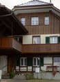 Two-story wooden house with green shutters, a wooden balcony, and a small porch area with a bench and potted plants.