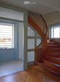 Spacious hallway with wooden flooring, a wooden staircase leading upwards, and large windows providing natural light. The walls are painted in neutral tones, and there is a small window seat with a few decorative items on the left side.