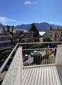 Balcony with metal railing overlooking a scenic view of mountains, a lake, and surrounding buildings. Several parked cars are visible below.