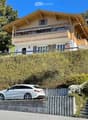 Two-story wooden house with balconies, surrounded by greenery, with a parked car in front.