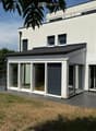 Modern two-story house with white exterior, large windows, and a raised ground floor. Features include a balcony on the upper level and a spacious patio area with glass doors leading to the interior.