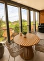 Balcony with a round wooden table, four white wicker chairs, a small potted plant, and a decorative vase. Large windows offer a scenic view of a countryside landscape.