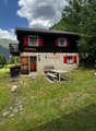 Traditional wooden chalet with red shutters, stone walls, and a balcony overlooking a mountain view. Outdoor seating area with a picnic table and benches.