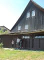 A two-story wooden house with a steep, dark wooden roof and multiple windows. The house has a covered entrance area with large windows and a door. The surrounding area includes a grassy lawn and some trees.