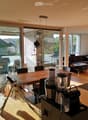 Open-plan living and dining area with wooden flooring, large windows, a sofa, a coffee table, a dining table with chairs, and a kitchen counter with a coffee machine and blender.