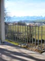 Balcony with metal railing, tiled floor, overlooking a green field and distant mountains.