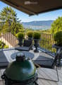 Balcony with wooden decking, black metal railing, and scenic mountain and lake view. Features a green grill, sofa, chairs, and potted plants.