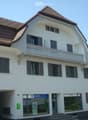 Building. A two-story white building with a brown tiled roof, multiple windows with shutters, and a small balcony on the upper floor. The ground floor features a glass entrance with a sign that reads "ZURICH".