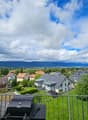 Balcony overlooking a suburban neighborhood with houses, green fields, and distant mountains under a partly cloudy sky. A grill is placed on the balcony.