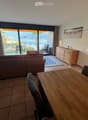 Bright living room with a view of the sea, featuring a sofa, dining table, chairs, and a cabinet.