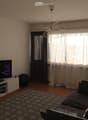 Modern living room with a grey sectional sofa, a flat-screen TV on a white stand, a patterned rug, and large windows with dark curtains. A decorative lamp hangs from the ceiling.