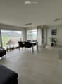 Open-plan living and dining area with large windows offering a scenic view, featuring a sofa, coffee table, dining table, chairs, and a modern kitchen area.