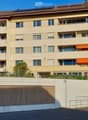 Four-story residential building with beige facade, multiple windows, balconies, and a fenced parking area at the base.
