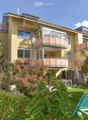 A three-story yellow building with multiple balconies and large windows, surrounded by greenery and a small patio area with outdoor furniture.