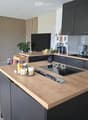 Modern kitchen with grey cabinets, wooden countertops, induction cooktop, sink, and various appliances. Includes a candle holder and a small plant on the counter.