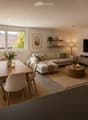 Modern living and dining area with a large window, a grey sectional sofa, a round coffee table, a dining table with chairs, a television, and decorative plants.